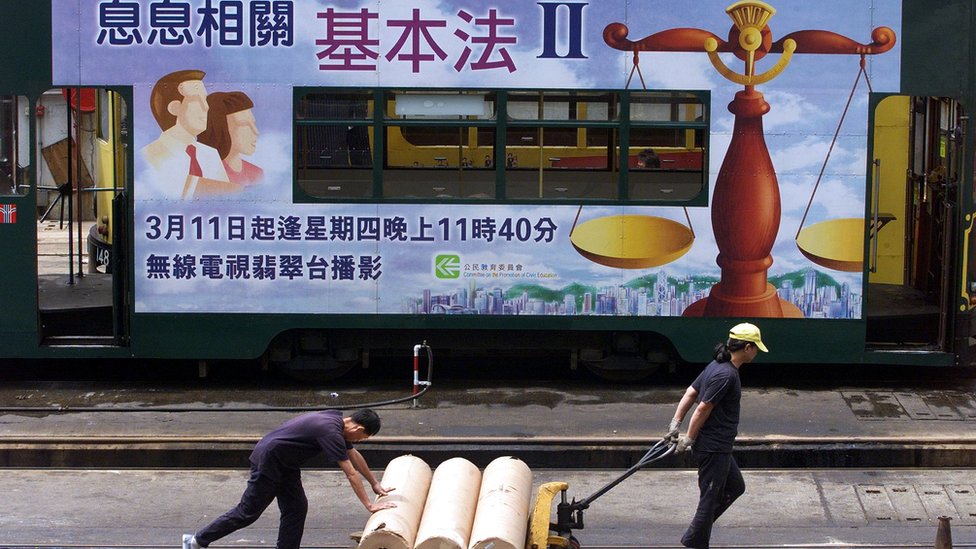Workers move supplies past a tram decorated with an advertisement encouraging Hong Kong people to increase their knowledge of the Basic Law, the territory's constitution, 07 May 1999 in Hong Kong. The government has been fiercely criticized by lawmakers for compremising the Basic Law and the independence of the judiciary by being non-committal when asked whether it would implement a high court ruling which gives the right of abode to some 1.67 million mainland born children of Hong Kong parents. The government has suggested that it may ask Beijing's national parliament to re-interpret the law, a move that would jeopardize Hong Kong's autonomy. (ELECTRONIC IMAGE) AFP PHOTO/Robyn BECK (Photo by ROBYN BECK / AFP) (Photo by ROBYN BECK/AFP via Getty Images)