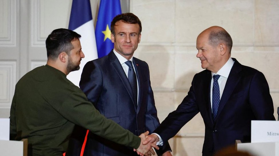 Volodymyr Zelensky shakes Olaf Scholz's hand while Emmanuel Macron looks on