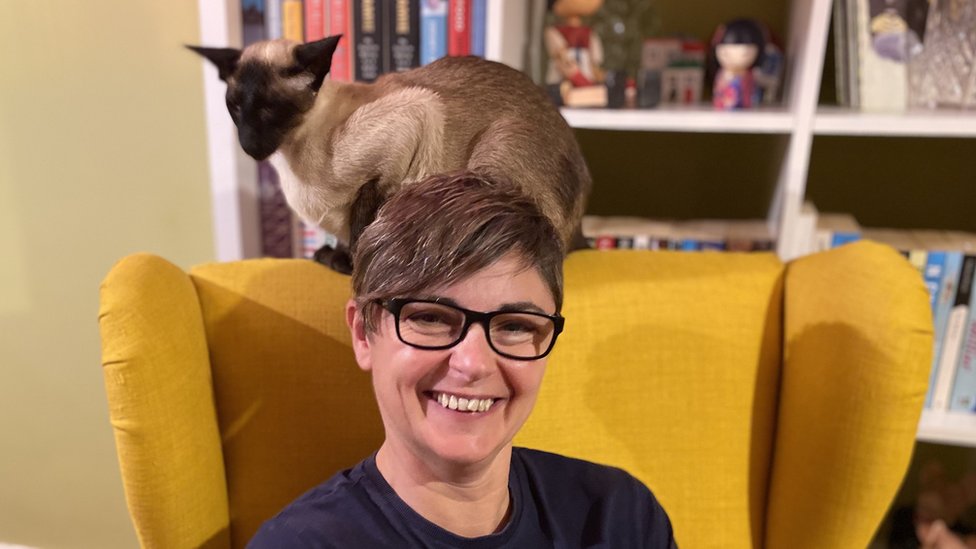 Louise Solomon sitting at home with a cat on her chair