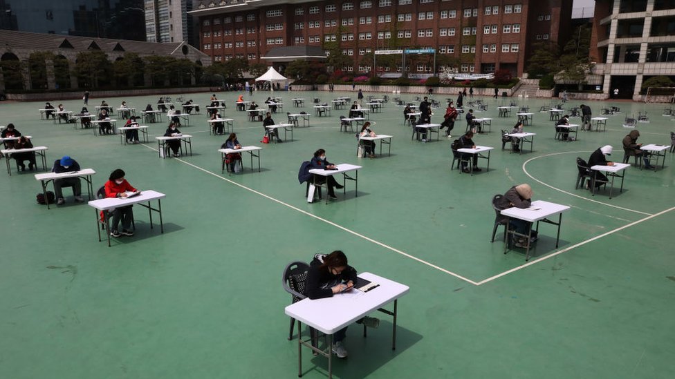 Южнокорейские студенты носят маски и сидят согласно правилам социального дистанцирования на экзамене по страхованию на детской площадке в университете Сеула в апреле 2020 года