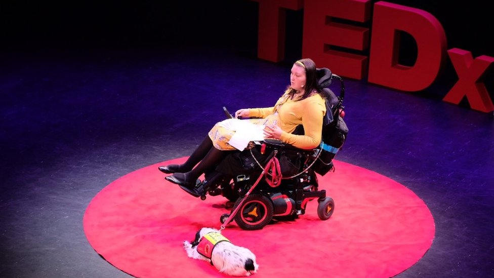 Lucy Watts speaking at a TedX event with her support dog Molly at her side