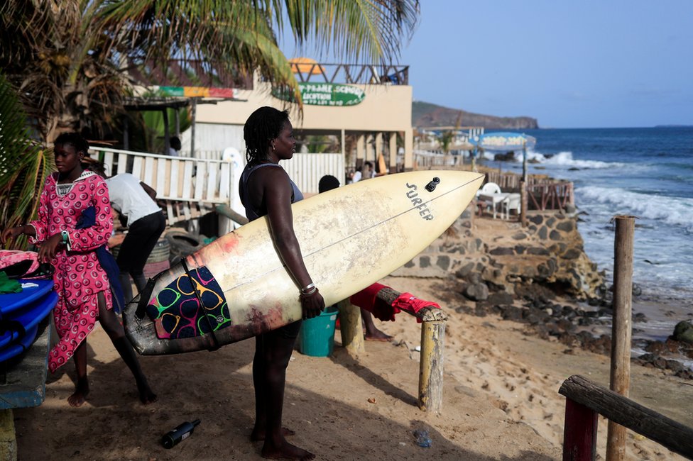 Khadjou Sambe stands on the beach with her surfboard and looks out to sea