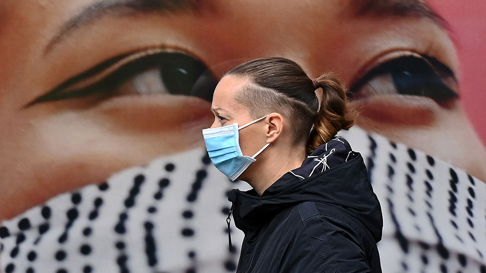 Pedestrian wearing mask in front of poster of woman wearing mask in West London