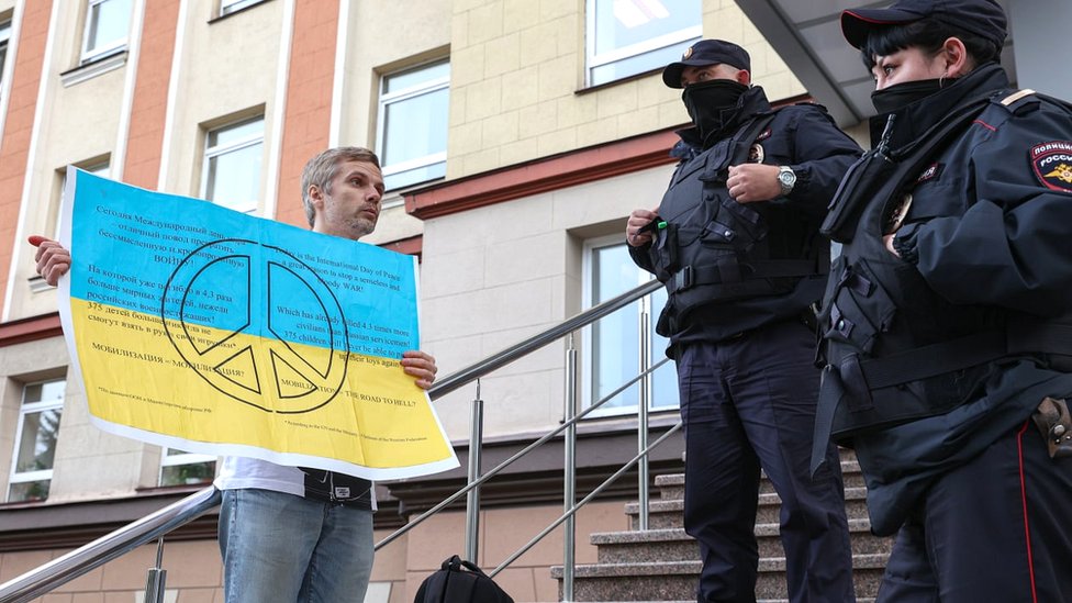 Un hombre con una pancarta con el simbolo de la paz y los colores de la bandera de Ucrania protesta frente a dos policías.