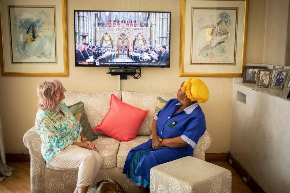 سيدتان تجلسان على أريكة في منزل وتنظران إلى شاشة التلفزيون التي تظهر موكب جنازة الملكة. ترتدي إحدى النساء زياً أزرق مع غطاء أصفر للرأس أصفر بينما ترتدي الأخرى بنطالاً أبيضاً وقميصاً أزرق اللون.