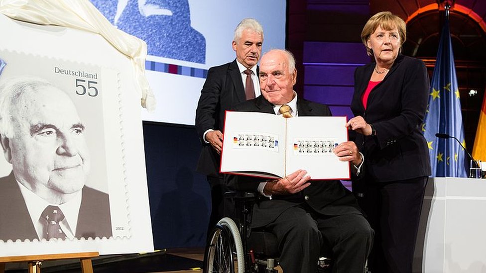 A 2012 picture of Angela Merkel presenting a commemorative postal stamp showing former German Chancellor Helmut Kohl as he looks on
