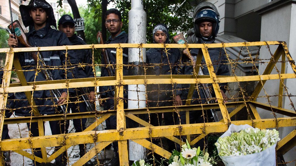 Бангладешские полицейские охраняют контрольно-пропускной пункт, где доброжелатели оставили цветочные композиции на дороге, ведущей к высококлассному кафе, которое было местом кровавой осады в Дакке, 3 июля 2016 г.