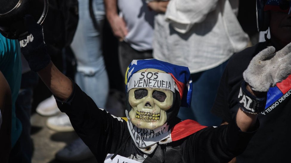Venezolano con una máscara de una calavera y un cártel que dice "SOS Venezuela".
