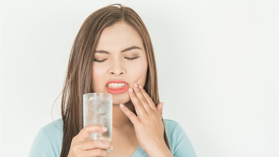 Una mujer con dolor de dientes, sosteniendo un vaso de agua y hielo.