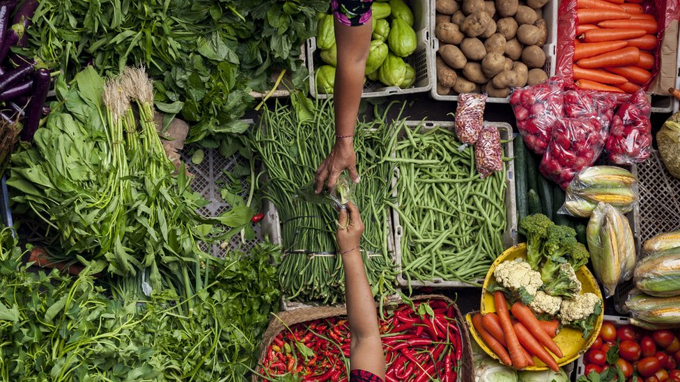 La mano de una mujer alcanza una bolsa con alimentos a otra, con un surtido de frutas y verduras en el fondo.