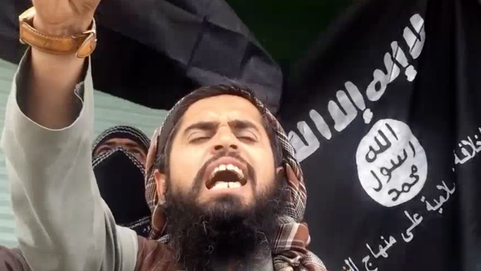 Скриншот видео ИГ, на котором мужчина кричит перед флагом ИГ
