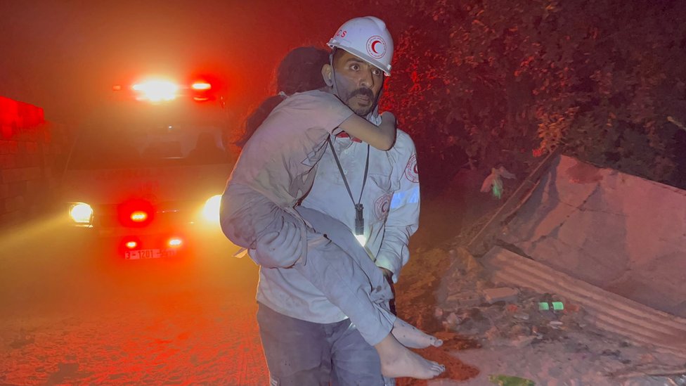 Paramedic carrying girl with red lights and rubble in background