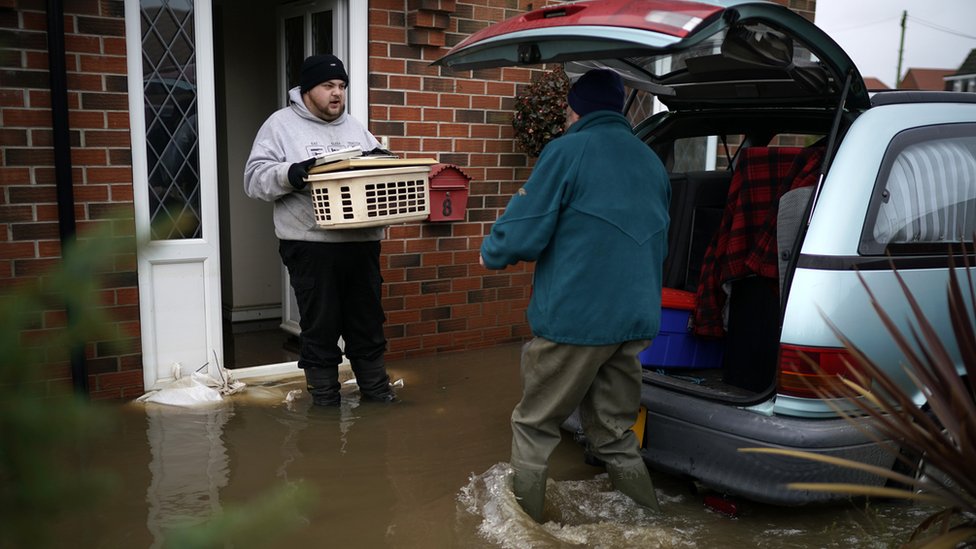 Двое мужчин выносят вещи из дома, пострадавшего от наводнения, и кладут его в багажник машины