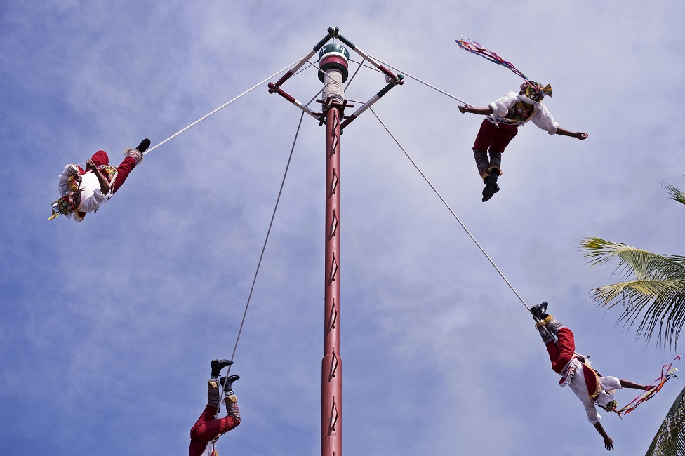Four men seeming to dance in the air, attached by ropes to a very tall pole