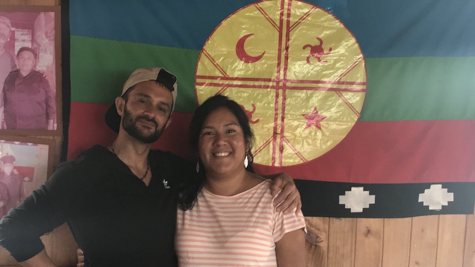 María y Daniel frente a una bandera mapuche.