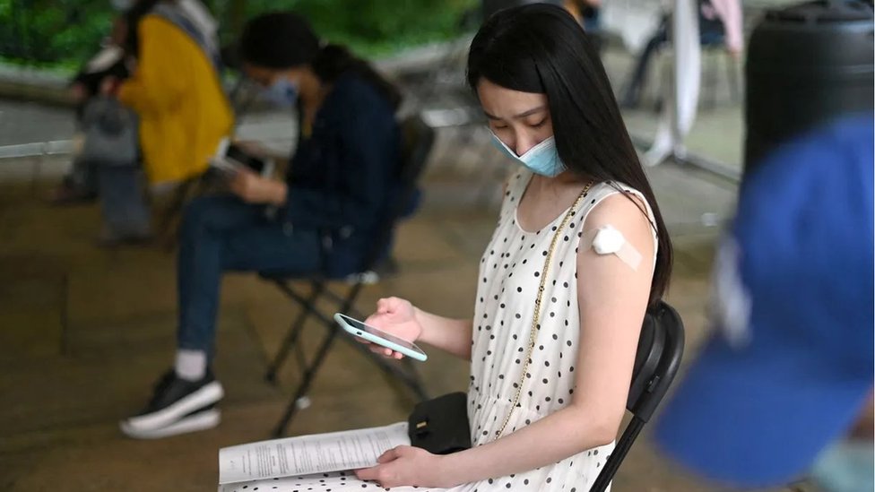 Jovem asiática sentada observa o celular, seu braço tem um curativo branco no local onde normalmente se toma vacinas, proximo ao ombro