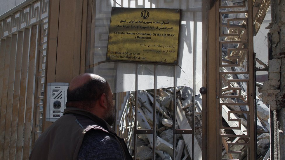 A man stands in front of the gates of the destroyed consular building
