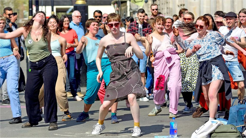 Grupo de pessoas brancas dança o que parece ser uma única coreografia na rua, em um dia ensolarado