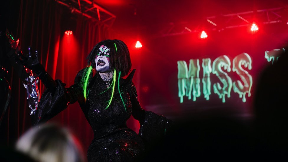 Danya no palco usando um vestido preto justo com maquiagem branca e extensões de cabelo neon