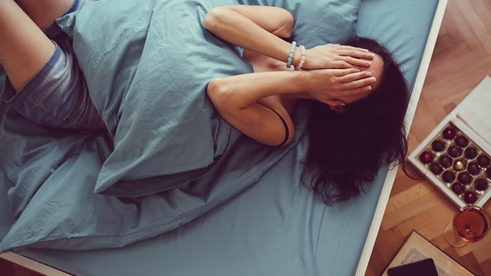 Una mujer echada en la cama, cubriéndose el rostro con las manos