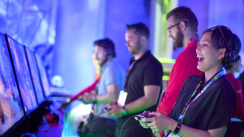 Четыре игрока на четырех игровых станциях держат контроллеры, а самый близкий человек громко смеется над невидимыми событиями на ее экране