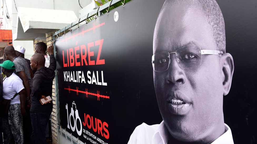 На этой файловой фотографии, сделанной 31 июля 2017 года, изображен баннер с фотографией мэра Дакара Халифы Салля, находящегося в тюрьме в ожидании суда по обвинению в хищении, которое, по словам сторонников, является политически мотивированным, и демонстрируется перед его офисом в Дакаре.