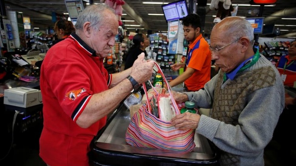 Un hombre empaca sus compras en una bolsa ayudado por otro