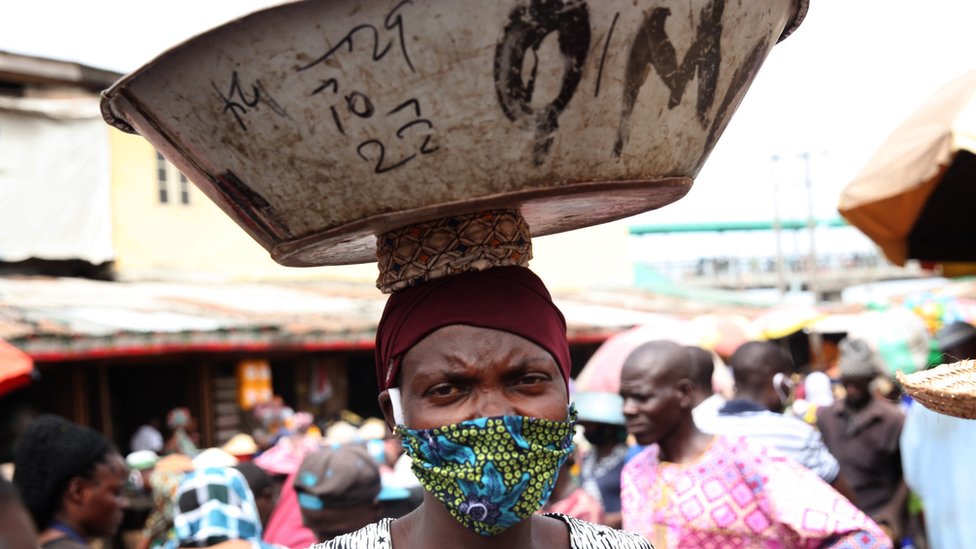 Женщина в маске для лица и несущая тарелку на голове в Лагосе, Нигерия - май 2020