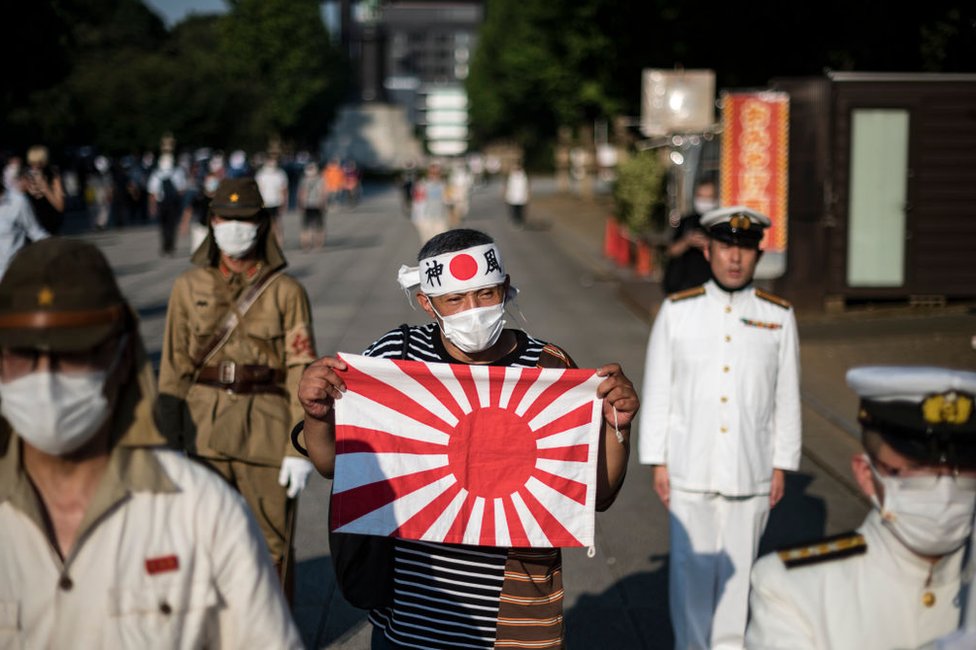 Un hombre sostiene la bandera del sol naciente entre otros que lucen uniformes del ejército imperial y la Marina durante un evento el 15 de agosto de 2020 en Tokio para conmemorar el 75 aniversario de la rendición de Japón en la Segunda Guerra Mundial.