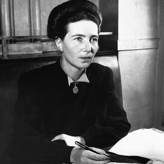 Simone de Beauvoir aparece em foto sentada e diante de uma mesa, com caneta na mão