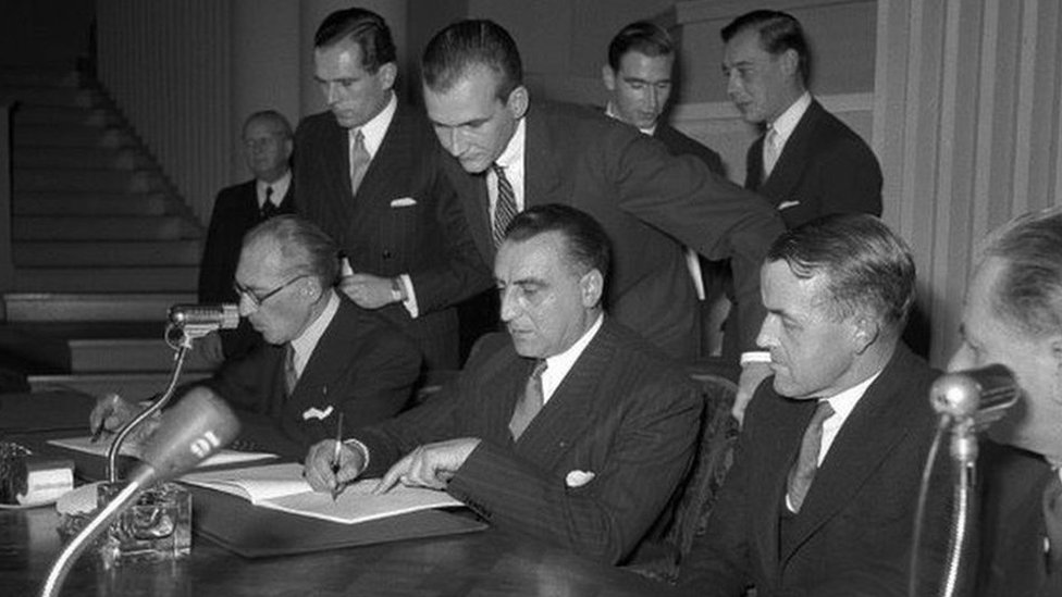 Black and white photo of a group of men gathered around a table signing a document