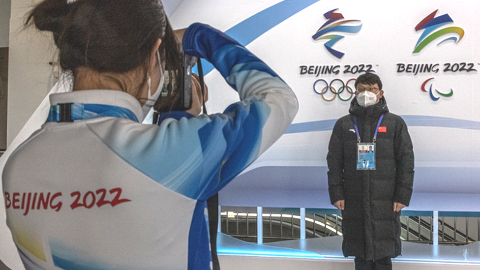 A woman snaps a photo of a man wearing a face mask next to the logos of the Beijing 2022 Olympic and Paralympic Winter Games inside the Olympic main media centre