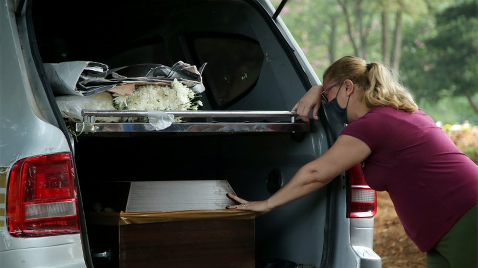 Mulher de m&aacute;scara chora encostada em carro com caix&atilde;o dentro