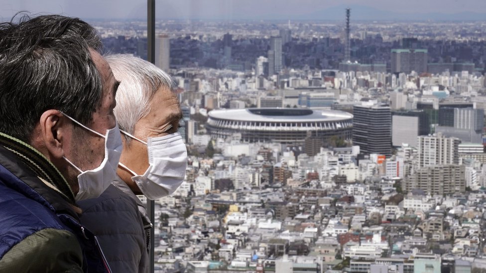 An elderly couple wearing protective face masks in front of the New National Stadium (background)