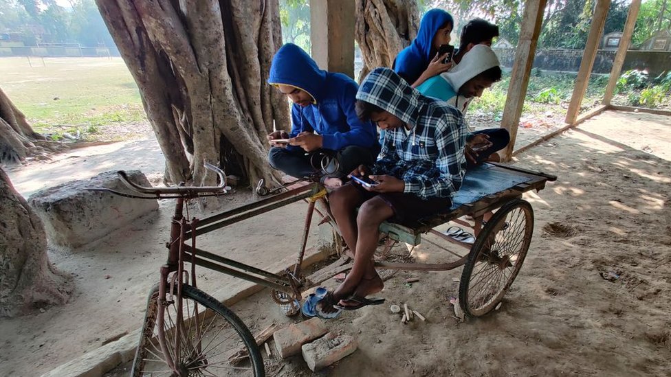 Children indulge in online gaming sitting on a broken down rickshaw.