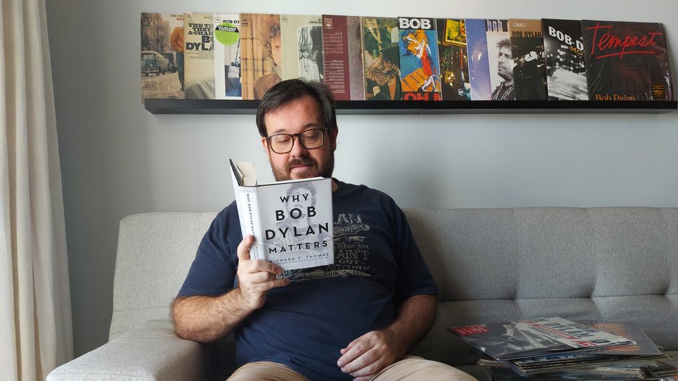 Pedro Couto sentado no sofá com livro sobre Bob Dylan na mão