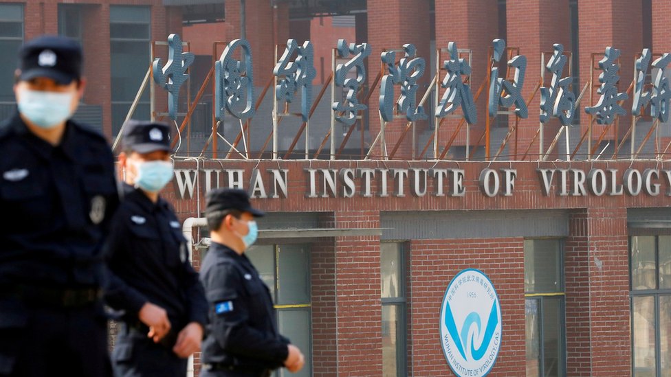Três agentes fardados e de máscara em frente a fachada com dizeres: Wuhan Institute of Virology