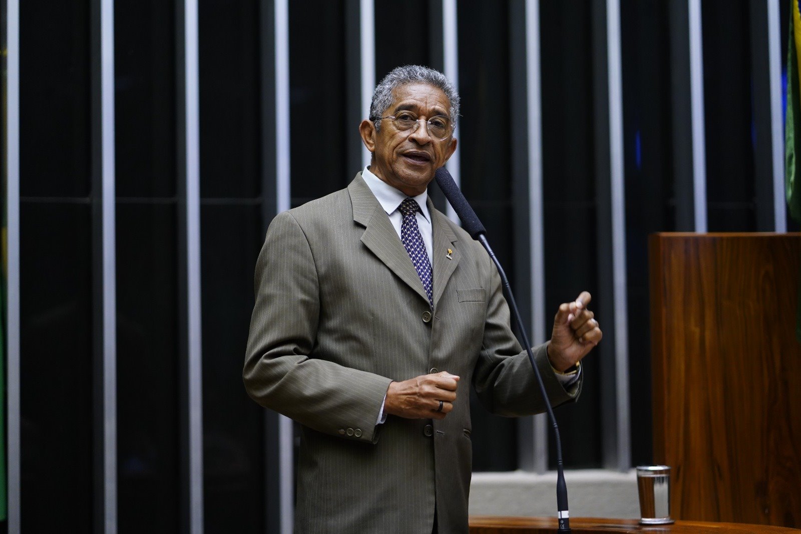 Retrato do deputado federal Vicentinho (PT-SP) na tribuna do Plenário da Câmara dos Deputados. Ele é um homem negro. Veste camisa social branca e púrpura com um terno cinza esverdeado.