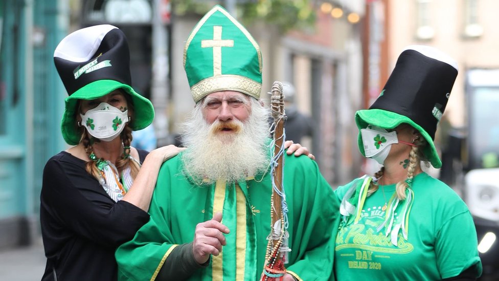 Две женщины в масках фотографируются с мужчиной в костюме Святого Патрика возле Temple Bar в Дублине