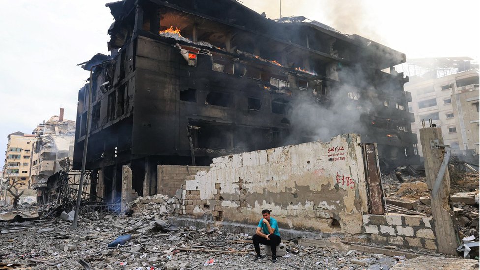 2023年10月10日，以色列對加沙市裏馬爾區發動空襲後，一名巴勒斯坦男子坐在一座燒焦的建築物前，大樓內部正熊熊燃燒。