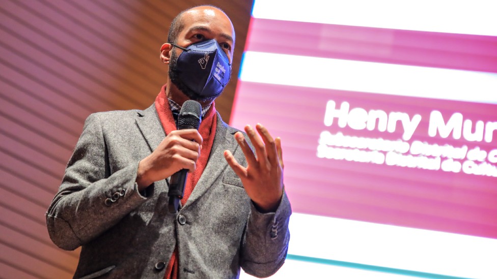 Henry Murrain fala em evento de máscara, segurando microfone e com telão ao fundo