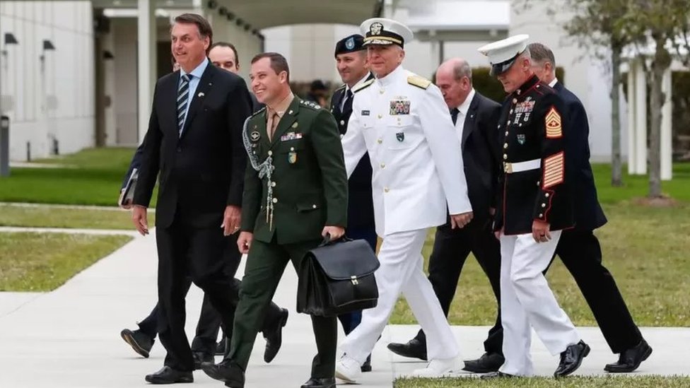 Homens idosos e um jovem, alguns de uniforme militar, caminham ao lado de Bolsonaro