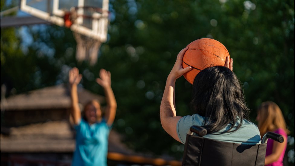 Criança de costas, em cadeira de rodas, jogando basquete