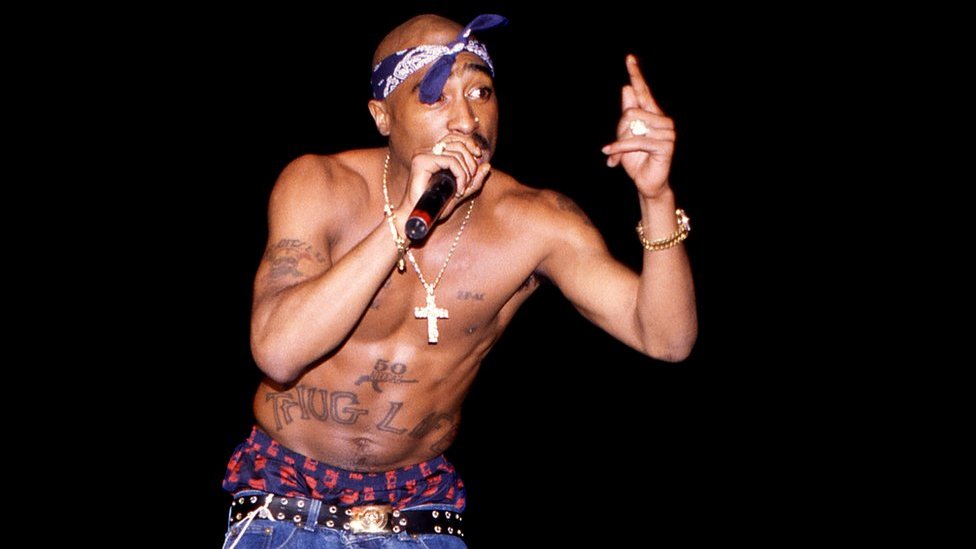Shirtless Tupac Shakur performing on stage with a microphone, wearing a blue bandana, gold jewelry, and jeans, with visible tattoos including "THUG LIFE" across his torso.