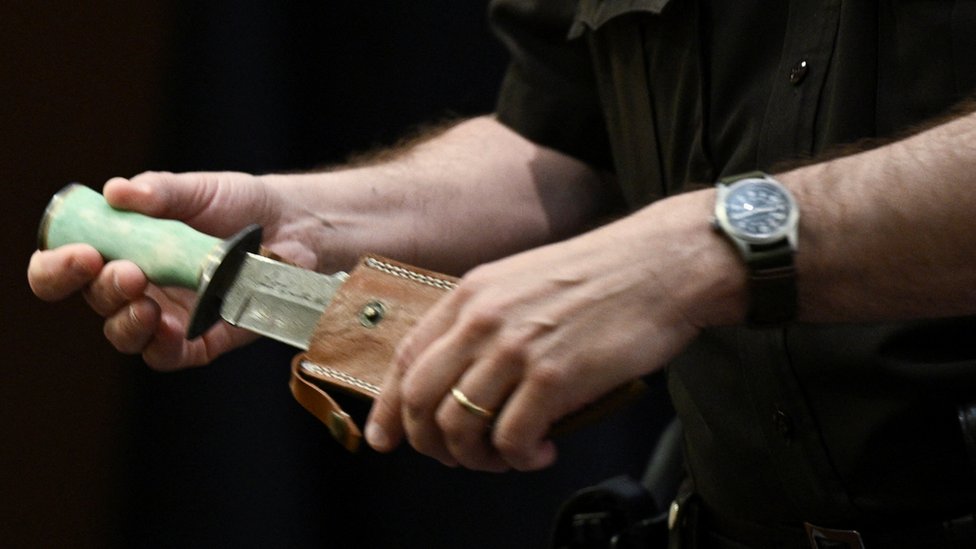 La manos de un alguacil mostrando un cuchillo de cazador con un mango verde y una funda de cuero.