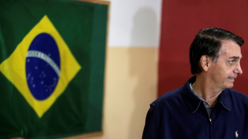 Bolsonaro em ambiente interno, olhando para o lado, perto de bandeira do Brasil