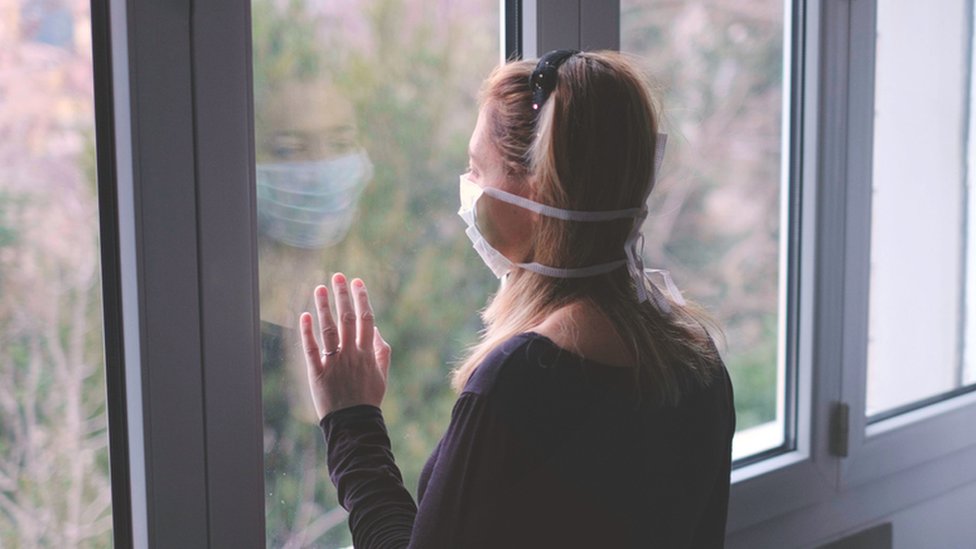 Woman in a mask looks at window with hand on the glass
