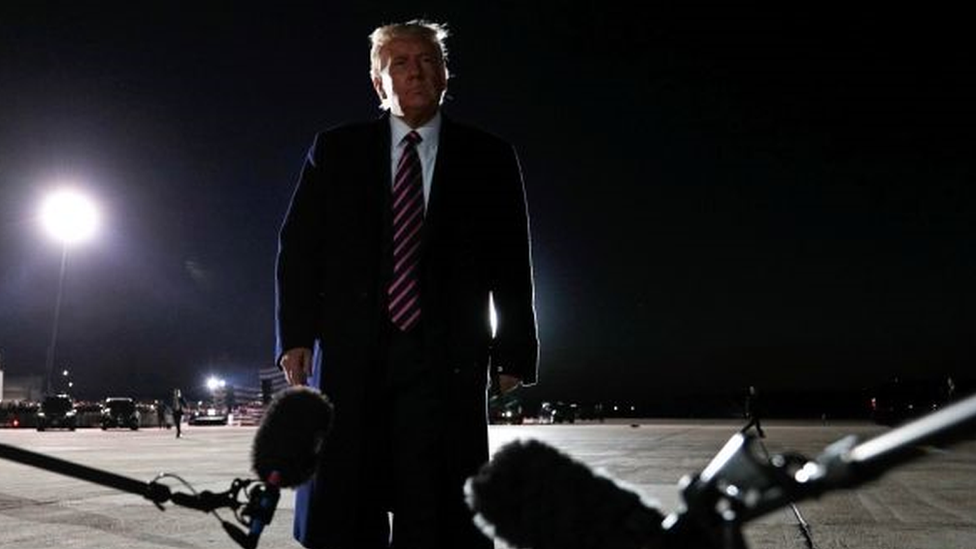 Trump em pé em pista, aparentemente de aeroporto, durante a noite e em frente a dois microfones