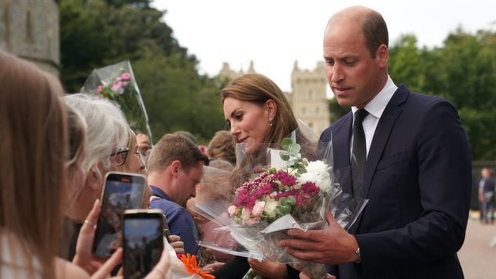 Foto colorida mostra o príncipe William, um homem branco adulto com cabelo rareando, e sua esposa Kate, uma mulher branca de cabelo castanho comprido, recebendo flores de diversas pessoas reunidas atrás de uma barreira