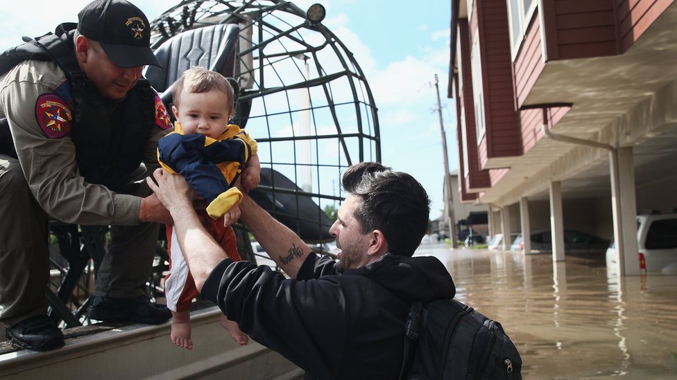 Un rescatista le entrega un bebé a un hombre que está hundido hasta la mitad del cuerpo por una inundación.